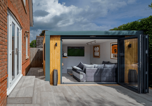 A garden room with sofas and a TV mounted on the wall. This man cave is framed by dark turquoise pillars and wood panelling. A zig-zag sliding  patio door stands opened.