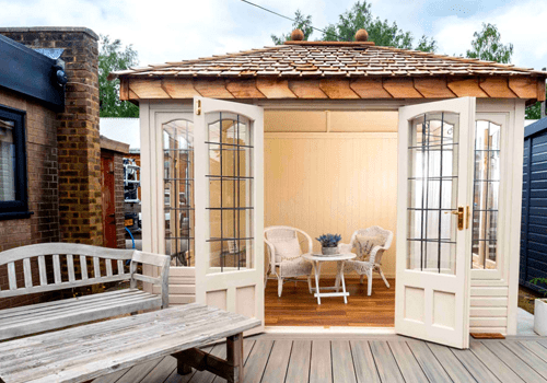 A small summer house with chairs and a table within. There is a warm light coming from within, and a long white bench and table outside on wooden decking.