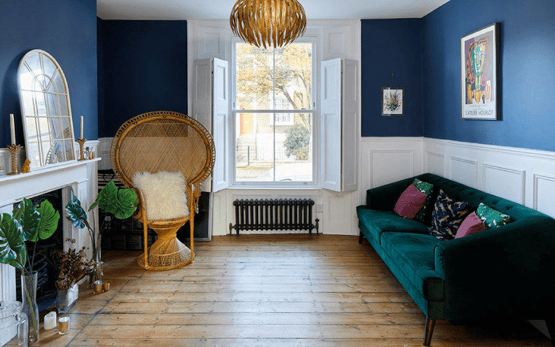 A blue-walled lounge space with a green sofa against one wall, opposite a fireplace. The floor is wood panelled and a wicker chair sits in the corner.